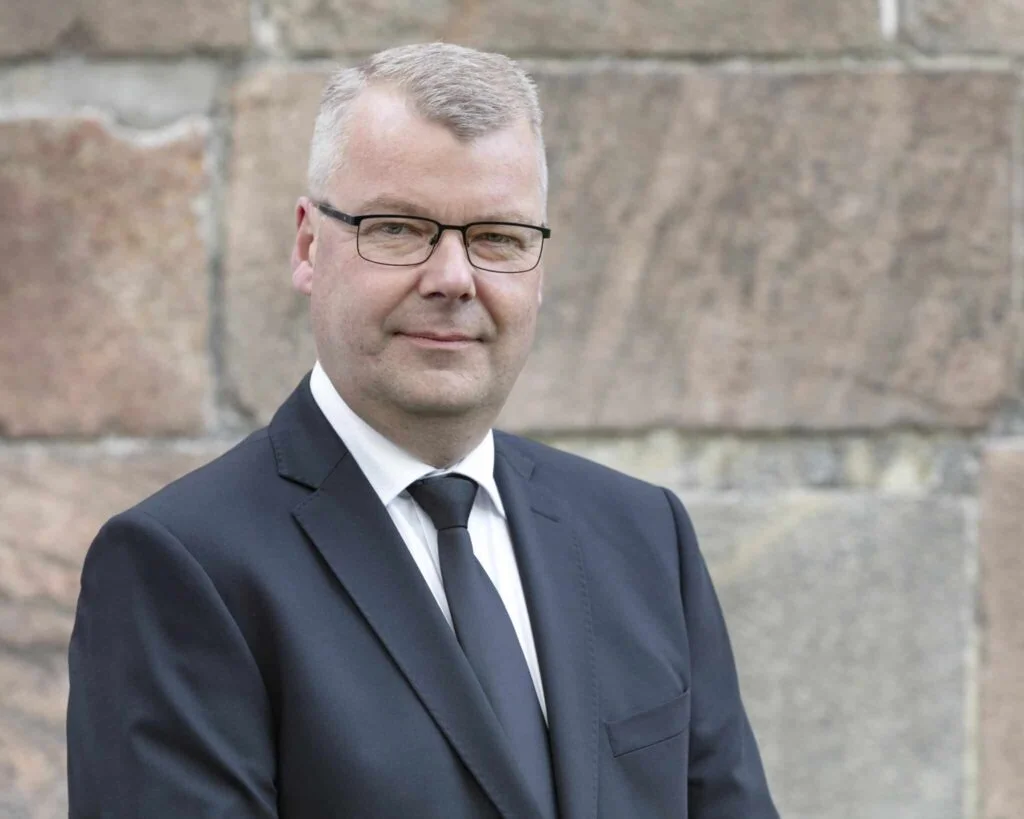 Mand i jakkesæt og briller foran stenvæg