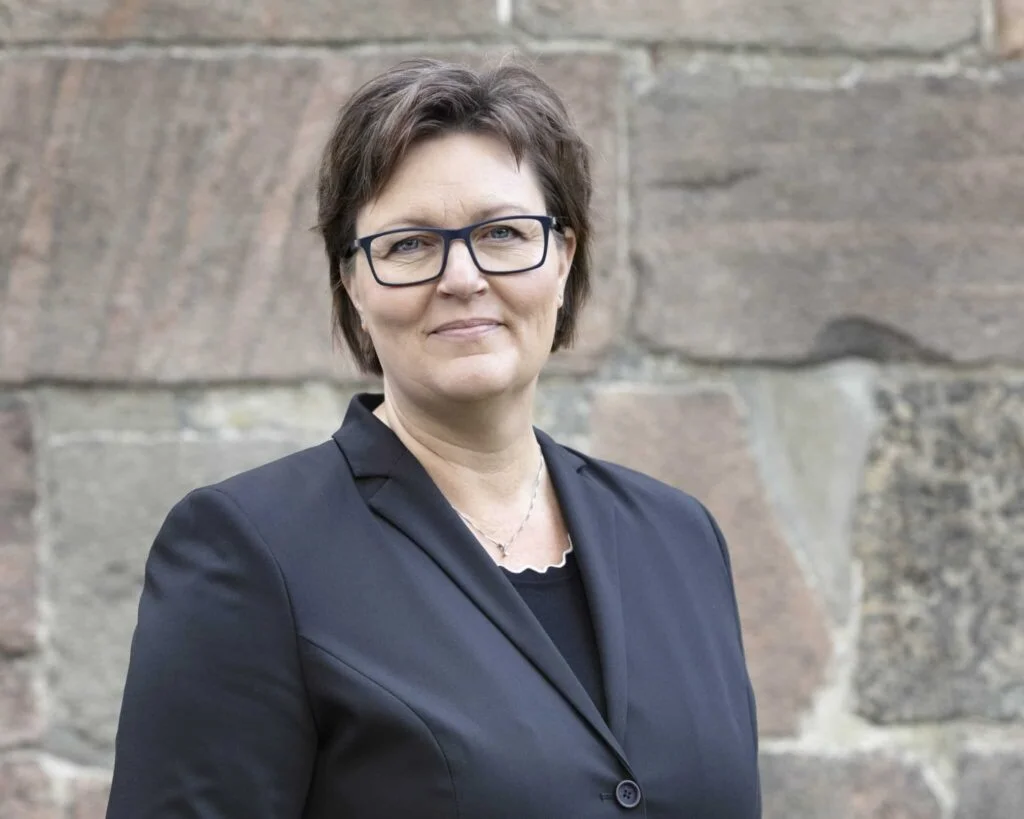 Kvinde i sort jakke med stenvæg i baggrunden.